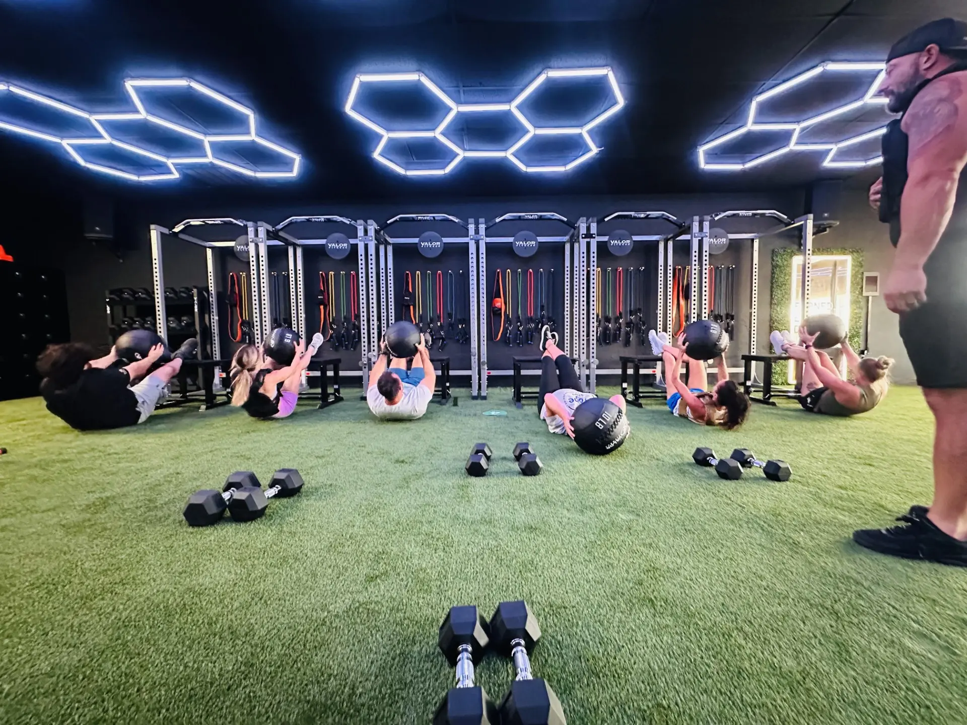 VestFit class doing medicine ball sit-ups on the turf floor with a coach watching.